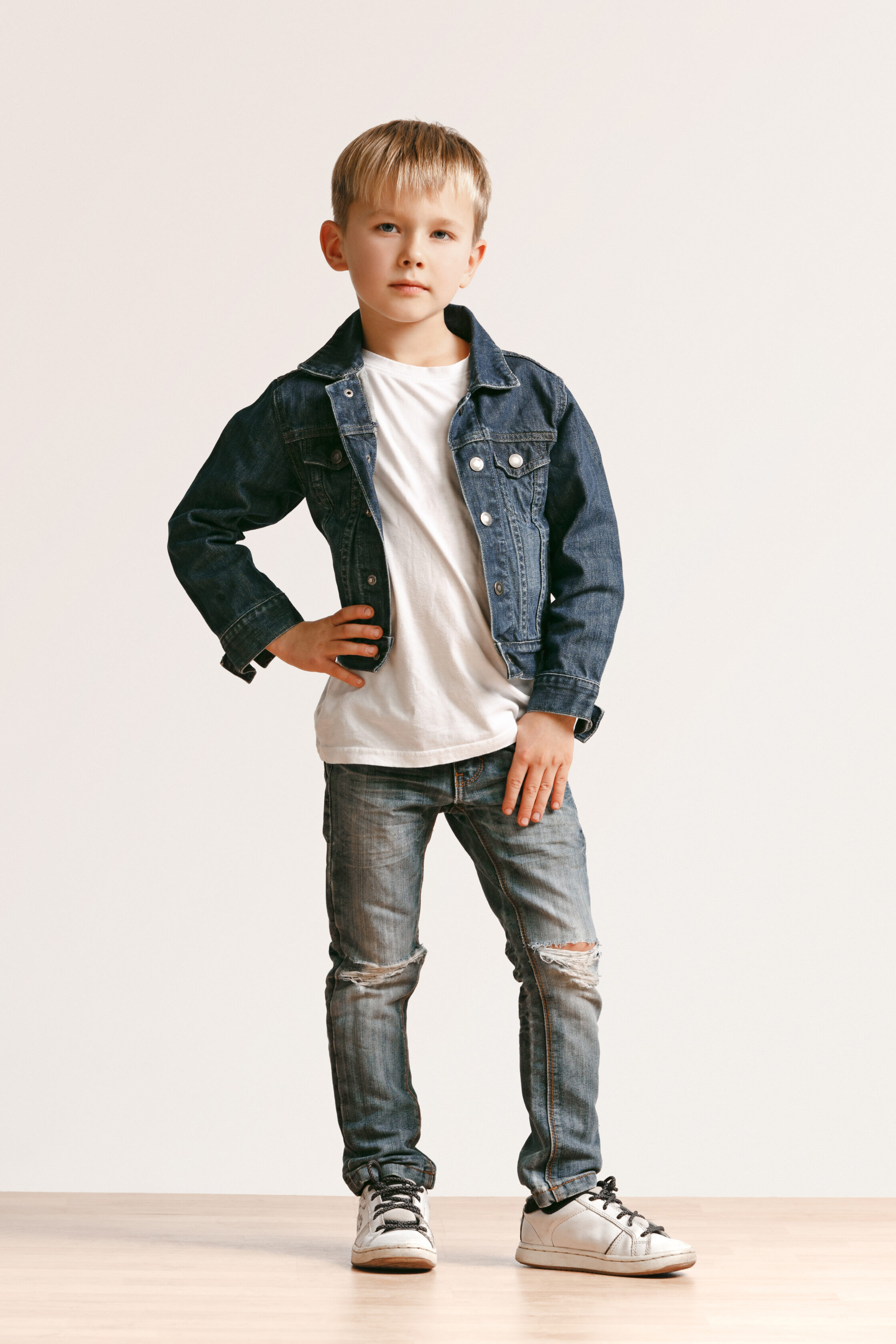 The portrait of cute little kid boy in stylish jeans clothes looking at camera against white studio wall. Kids fashion concept